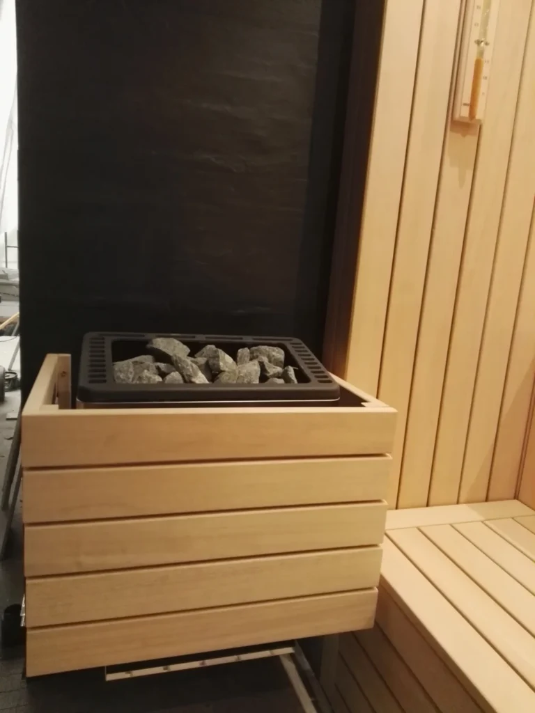 piec w saunie fińskiej z kamieniami, jasną drewnianą obudową i ławką obok